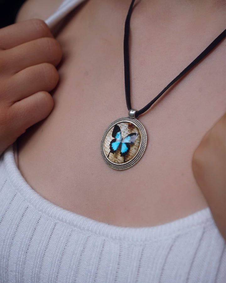 Bright blue Ulysses butterfly necklace on black cord with round silver-tone pendant worn with a white top