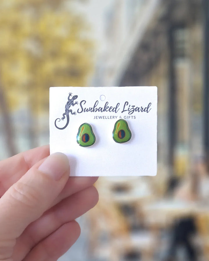 Avocado-themed earrings on a display card held by a hand with a blurred outdoor cafe background.