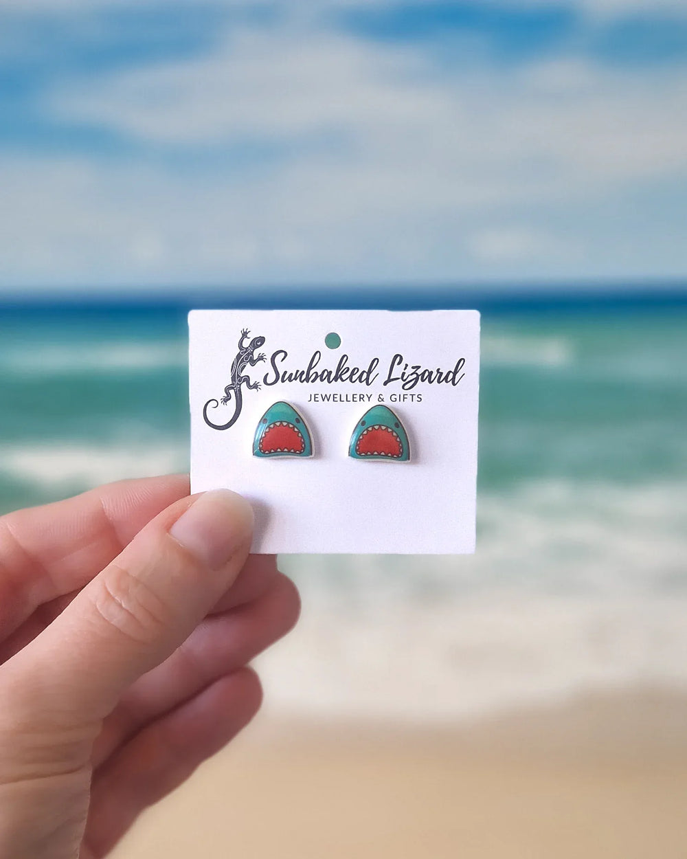 Shark Studs on earring card with held in front of a beach background