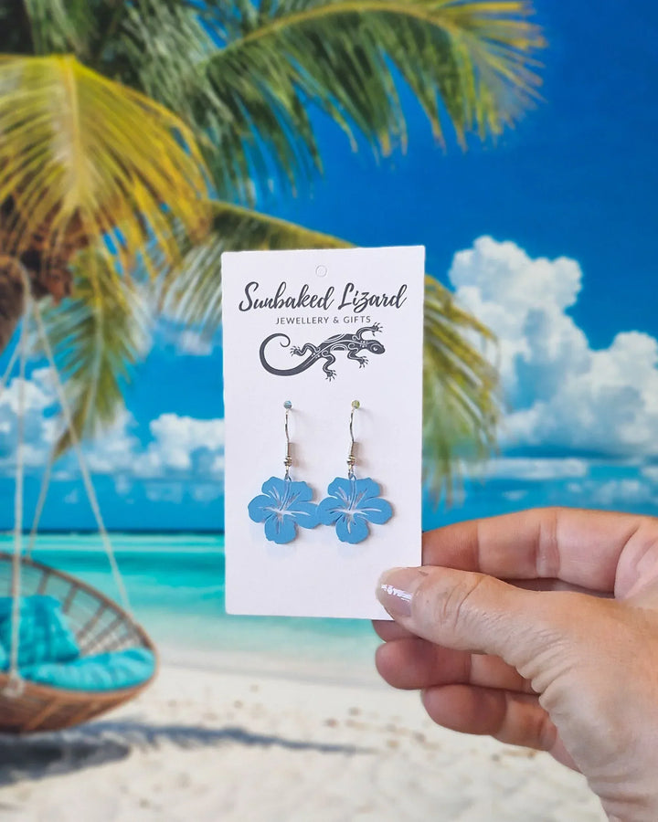 Filigree hibiscus drop earrings displayed on Sunbaked Lizard card against a tropical beach background