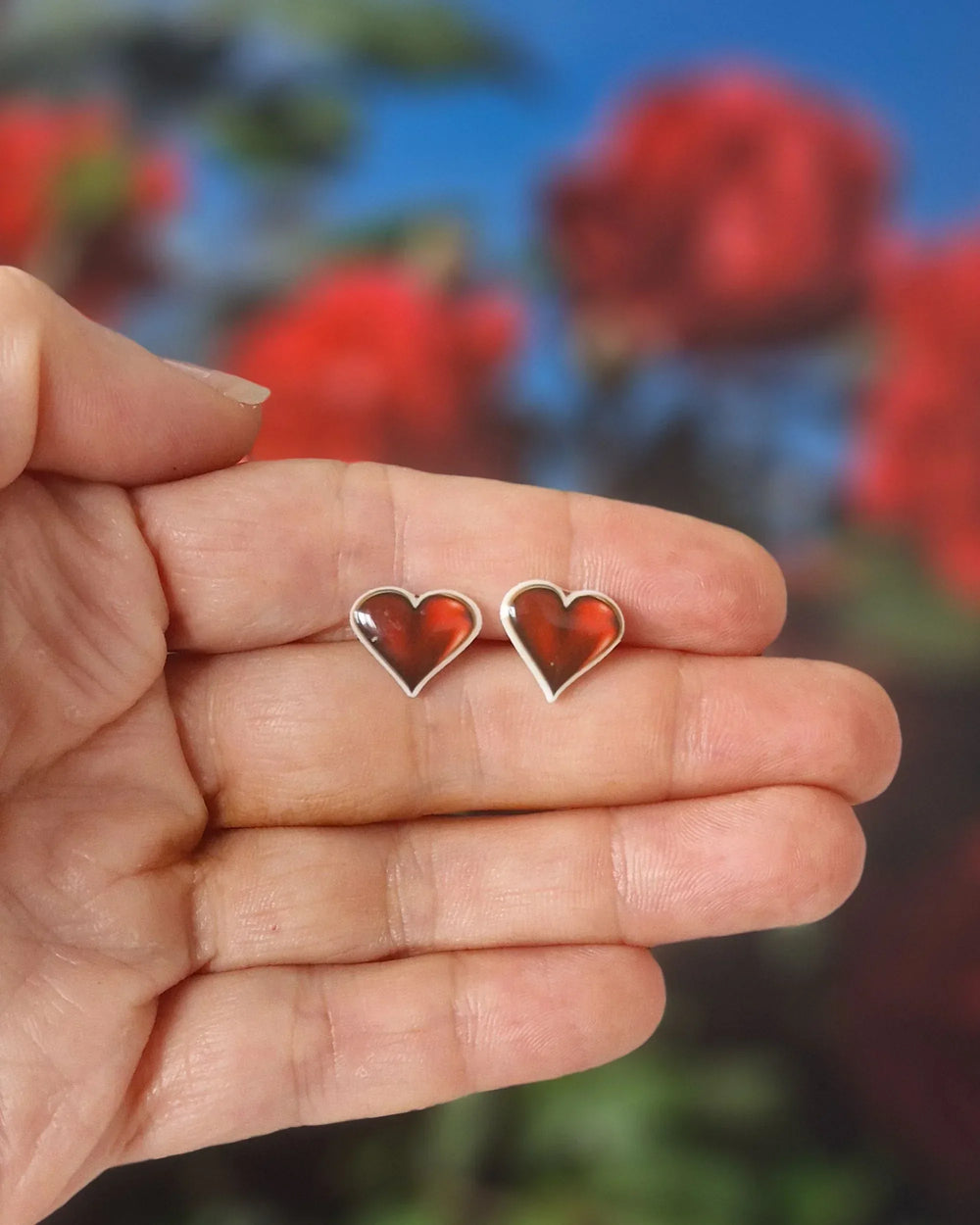 Shiny red heart stud earrings held in hand, showing glossy finish and white outline