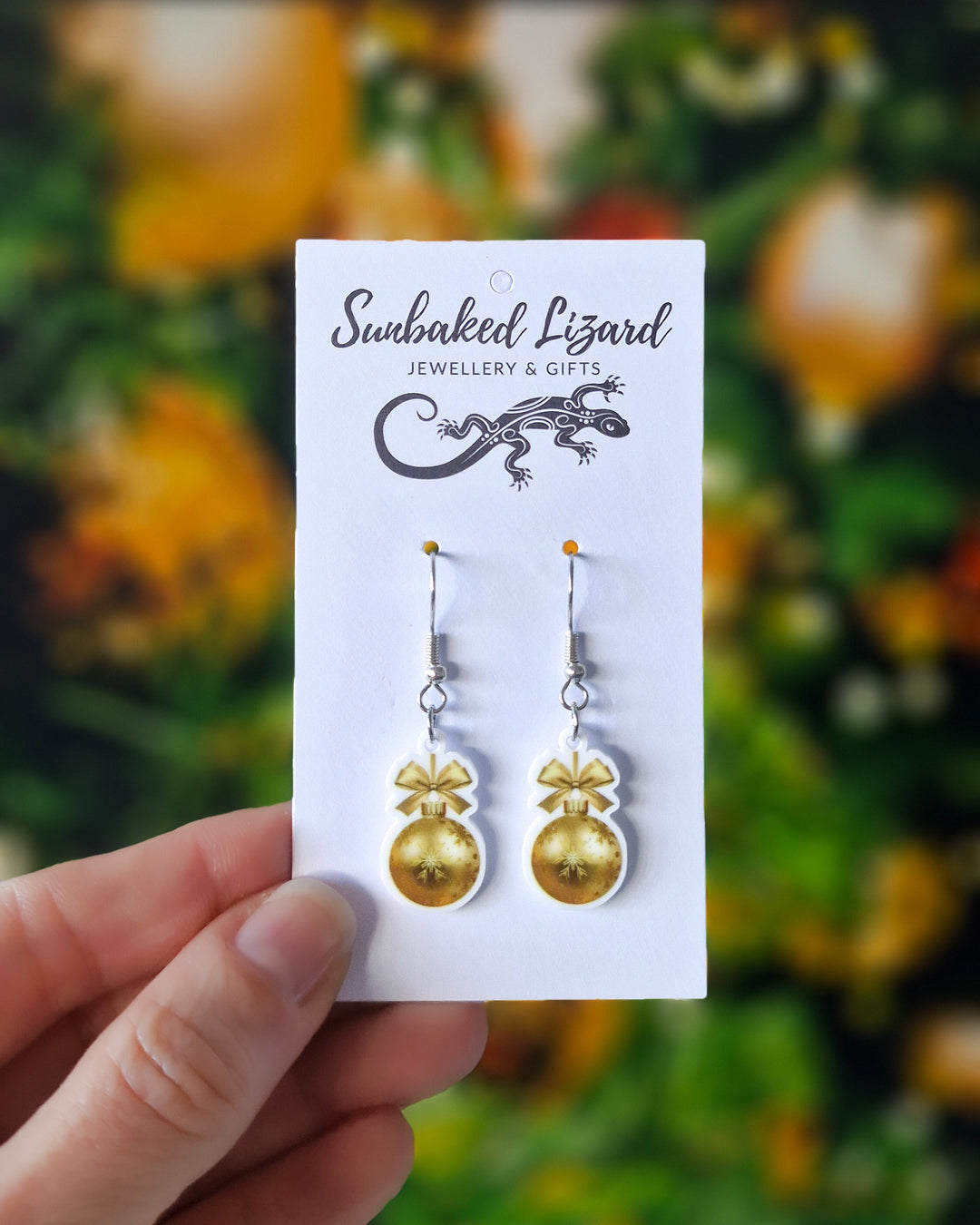 2D gold Christmas bauble earrings with a gold bow and snowflake print, hanging from silver hooks on a Sunbaked Lizard backing card to show scale