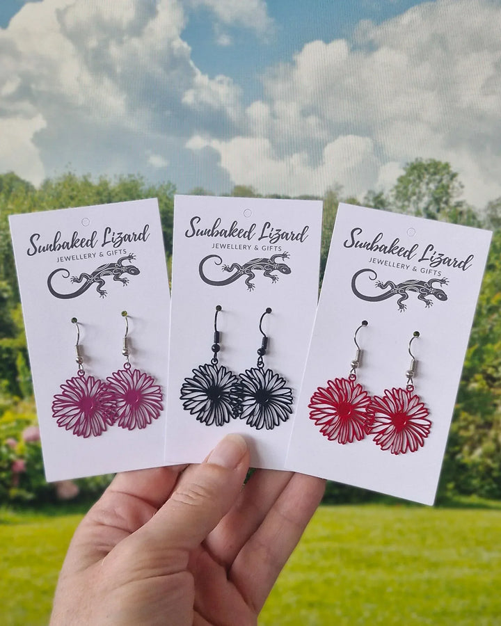 Filigree daisy drop earrings in pink, black and red displayed on Sunbaked Lizard jewellery cards outdoors