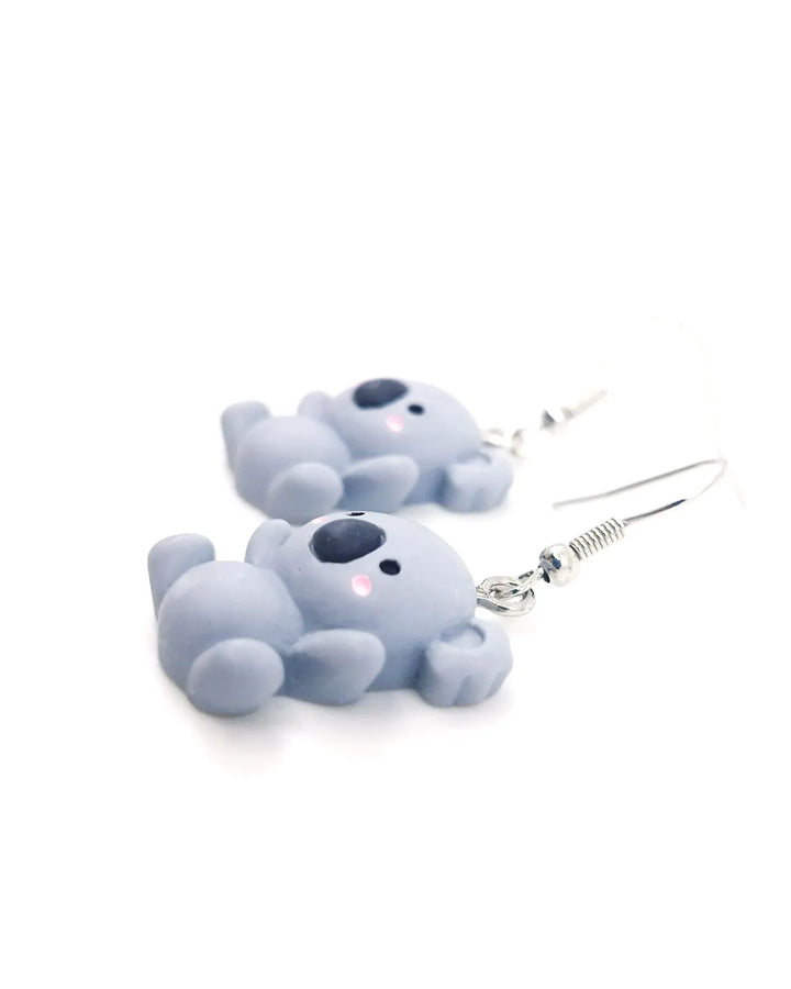 Angled close-up of cute grey koala dangle earrings with pink cheeks and silver hooks on a white background.
