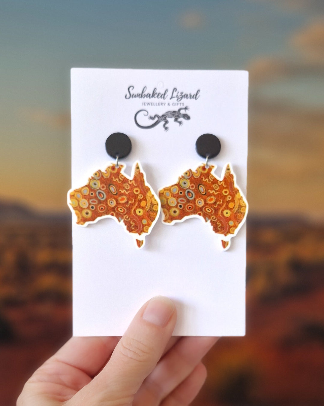 Orange Dreamscape Australia map earrings featuring swirling dot-style design. On a Sunbaked Lizard Jewellery &amp; Gifts card held against a blurred desert background
