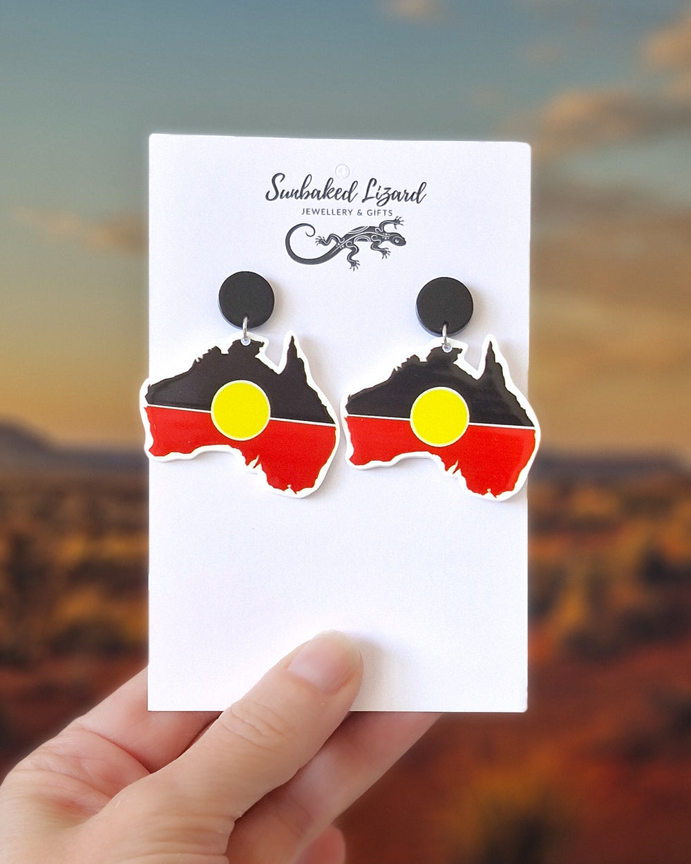 Australia map earrings with Indigenous inspired black, red and yellow colour design on a white card held in front of a desert background.