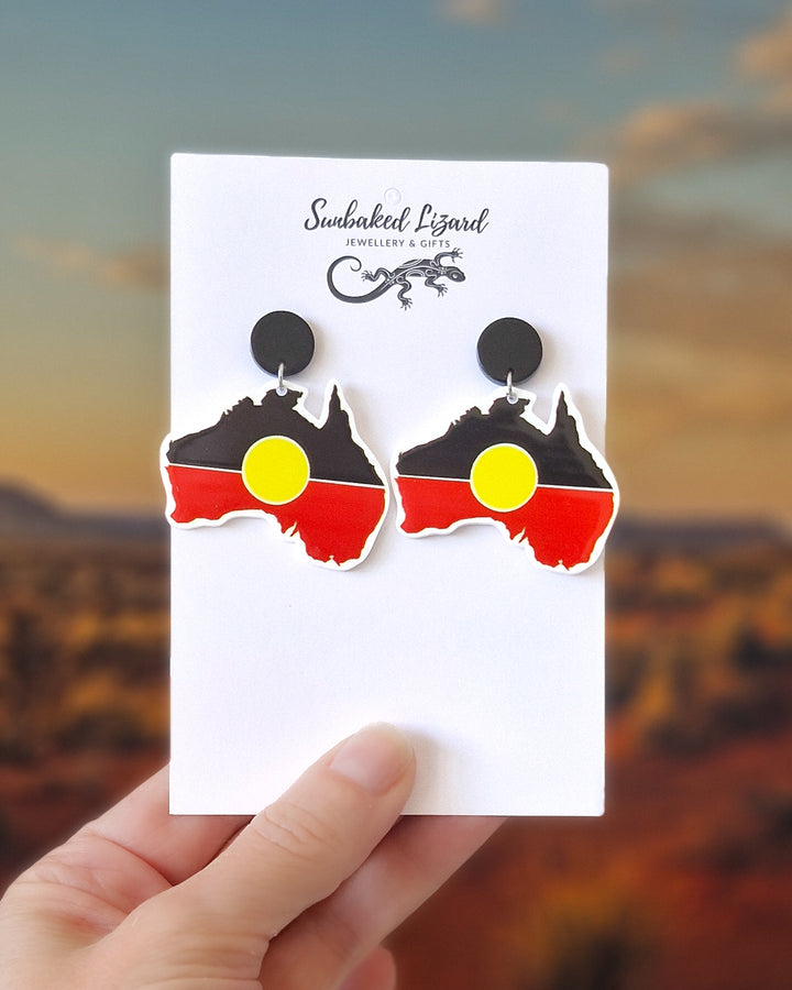 Australia map earrings with Indigenous inspired black, red and yellow colour design on a white card held in front of a desert background.