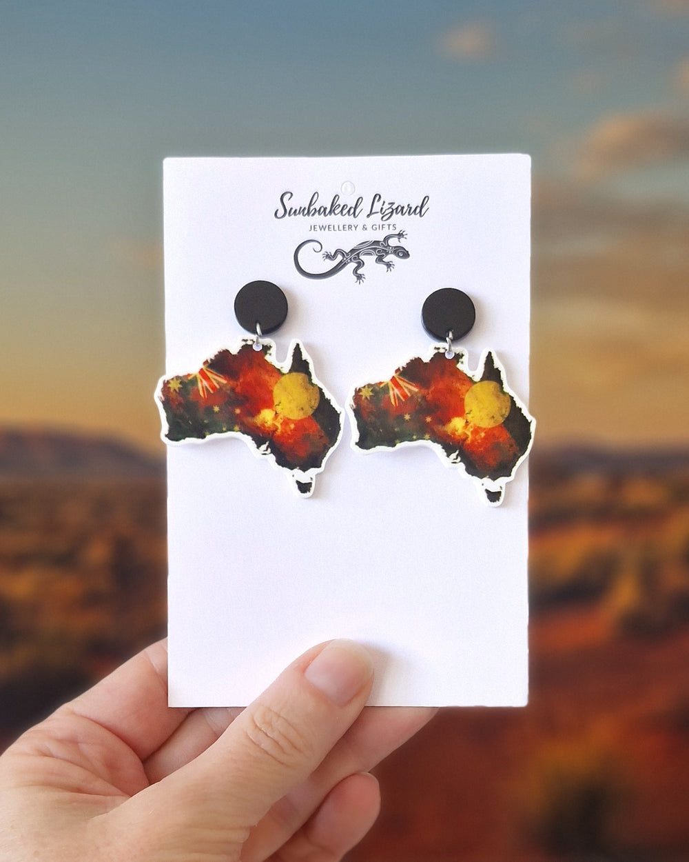 Australia map earrings featuring Indigenous inspired colours and Australian flag elements on a white card held in front of a desert background.