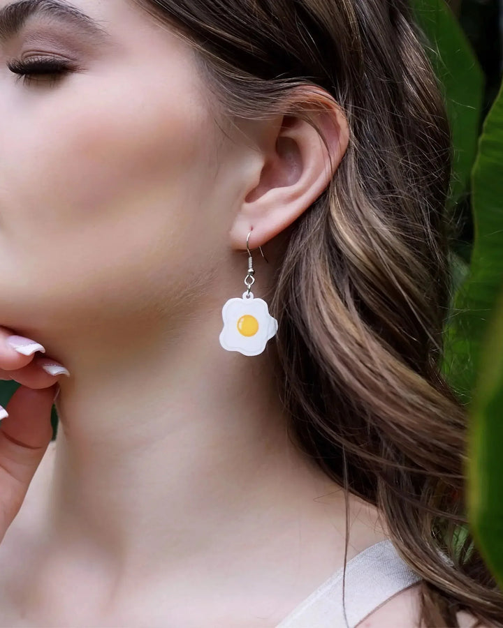 Side view of woman wearing a fried egg earring with greenery in the background