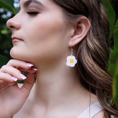 Side view of woman wearing a fried egg earring with greenery in the background