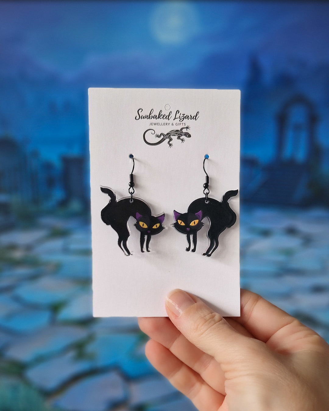 Hand holding Black cat dangle earrings with arched backs, yellow eyes and black hooks on a Sunbaked Lizard jewellery card to show scale