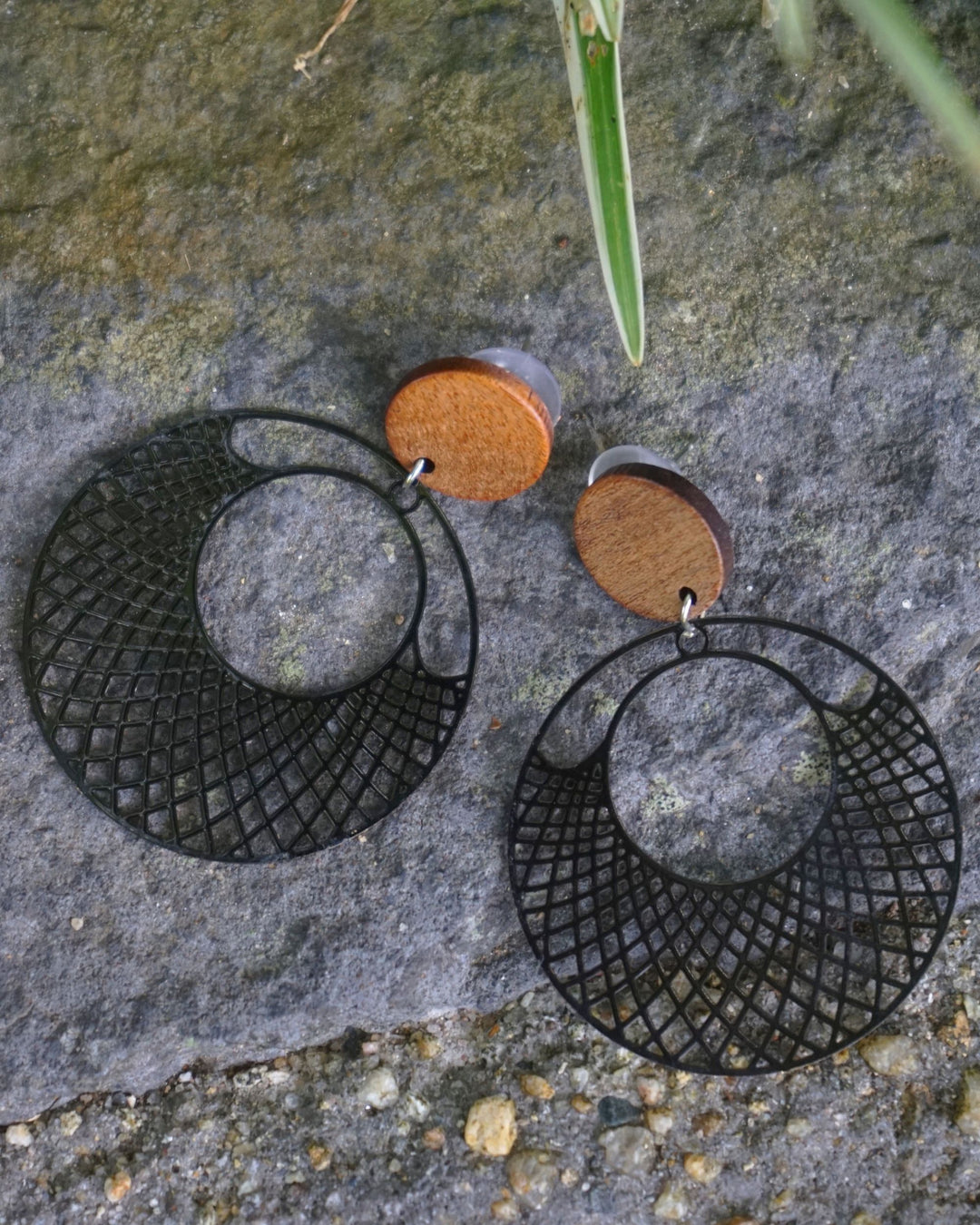Black filigree round earrings with timber stud top and cut-out lattice circle drop on a natural stone background.