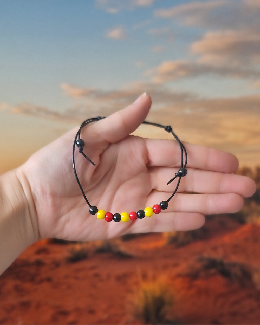 Hand holding Black Leather Indigenous, Red, Yellow & Black Beaded Anklet in front of a red desert background