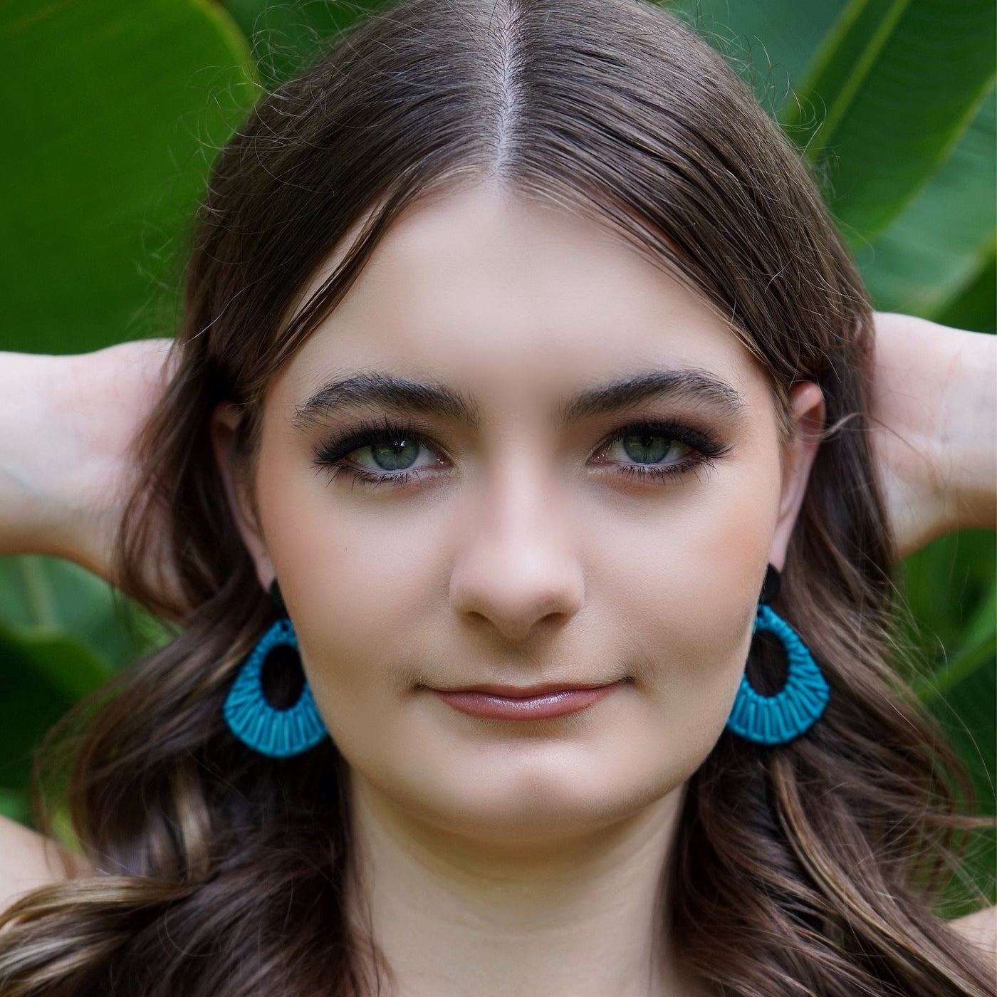 Front view of Woman with long brown hair and blue earrings against a green leafy background