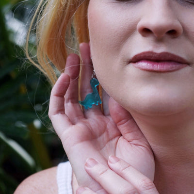Woman wearing blue dinosaur earrings with a blurred green background