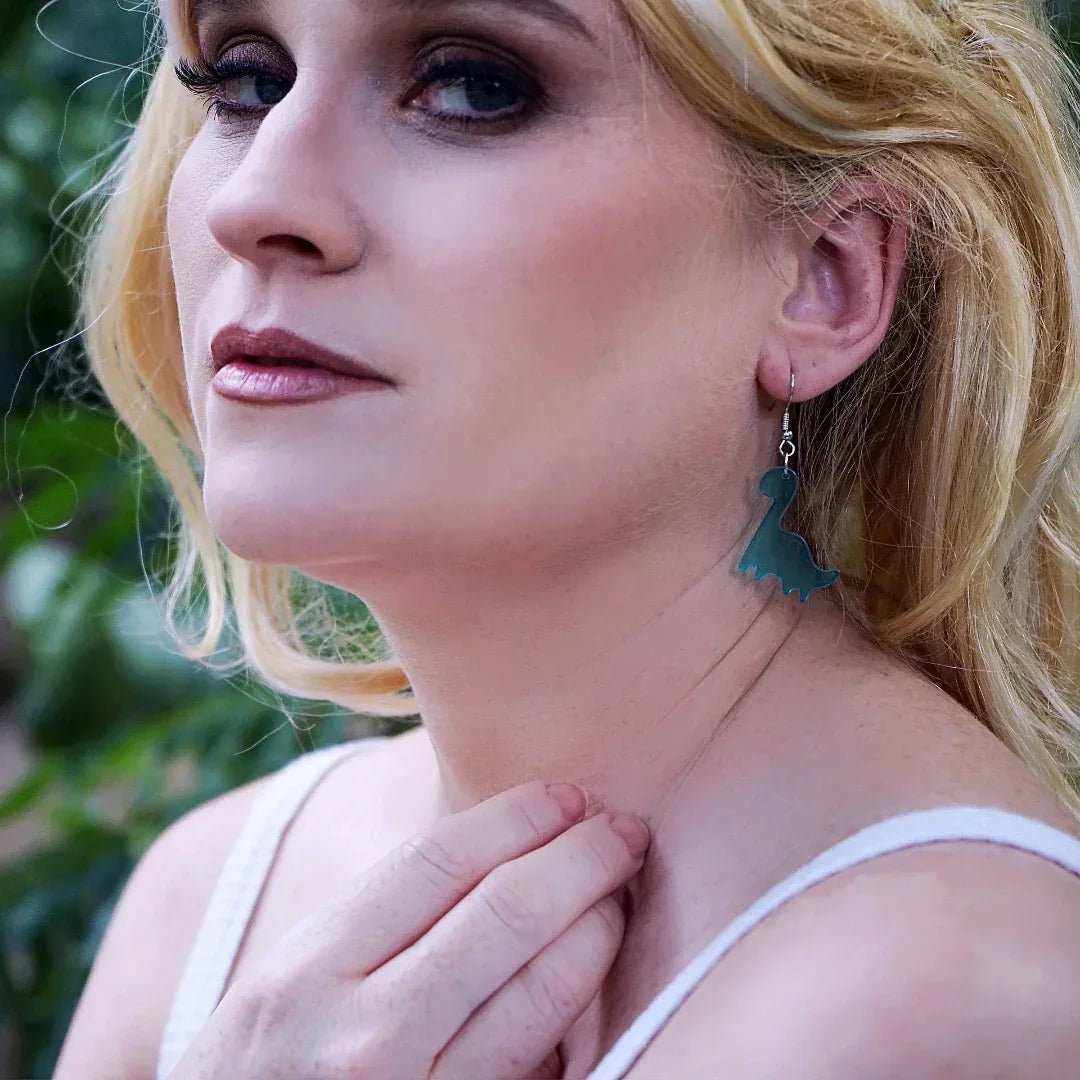 Woman with blonde hair and blue dinosaur earrings standing in a natural setting