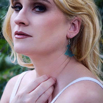 Woman with blonde hair and blue dinosaur earrings standing in a natural setting