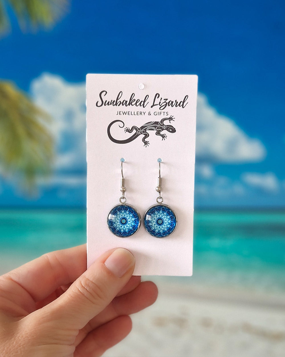 Hand holding vibrant Blue Mandala Pattern Drops on a white display card in front of a tropical beach background.