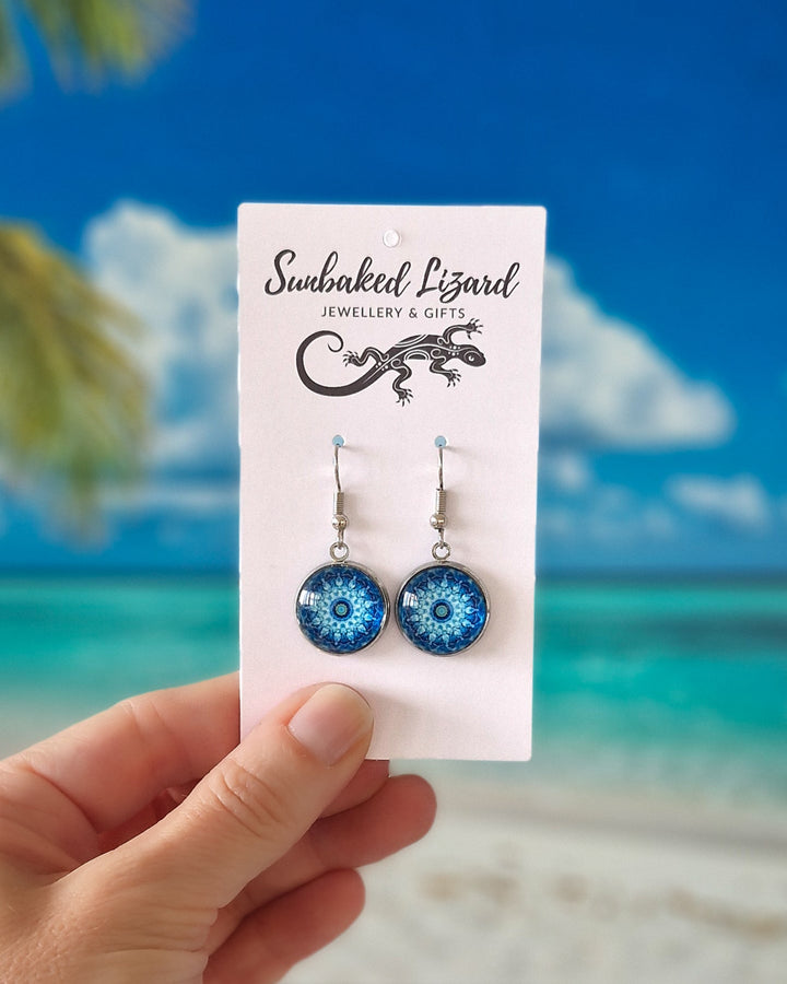 Hand holding vibrant Blue Mandala Pattern Drops on a white display card in front of a tropical beach background.