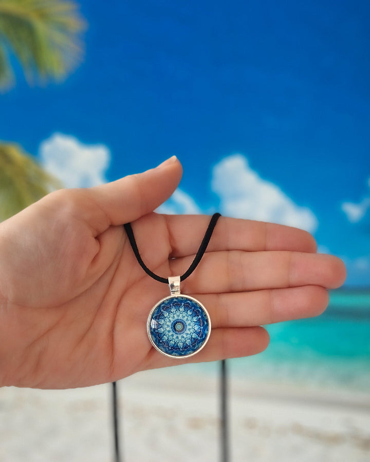 Hand holding necklace with a blue and silver mandala pendant with a tropical beach in the background