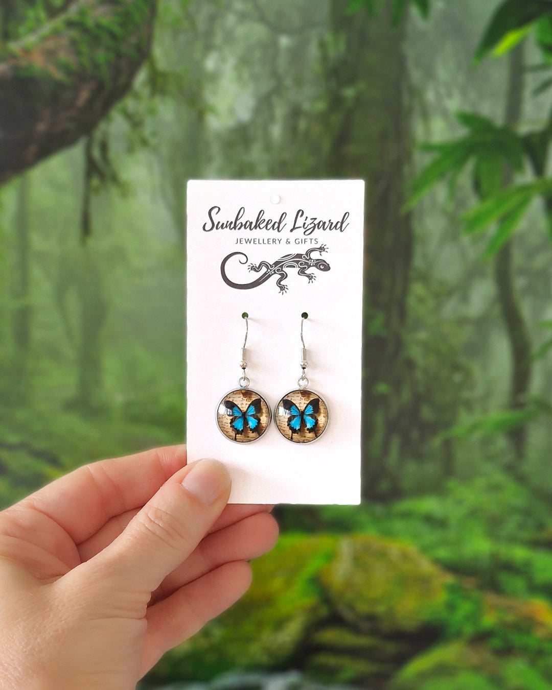 Hand holding vibrant Blue Ulysses Butterfly earrings on a display card with  a tropical rainforest background.