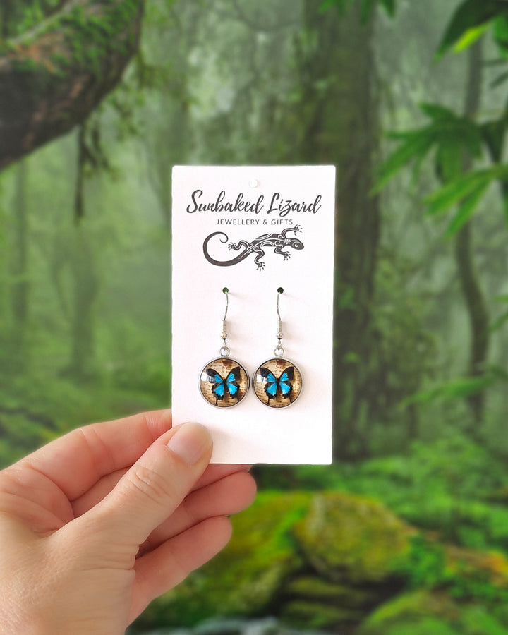 Hand holding vibrant Blue Ulysses Butterfly earrings on a display card with  a tropical rainforest background.