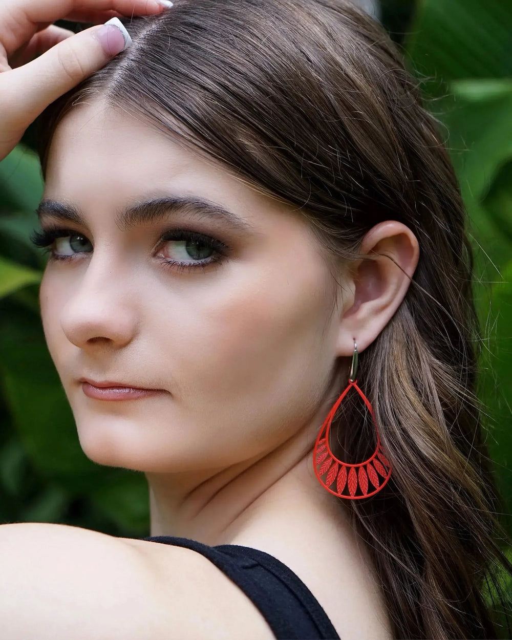 Model wearing bright orange Leafy Droplet Filigree Drop Earrings against green foliage