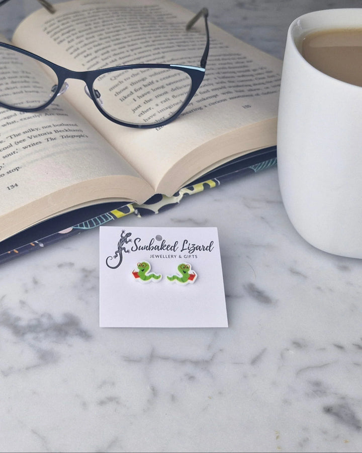 Open book with glasses on a marble table next to a mug of coffee and a pair of bookworm earrings.