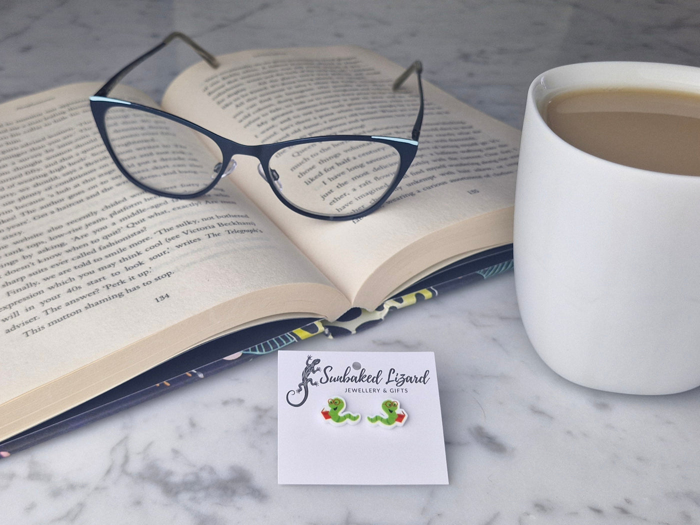 Open book with glasses on a marble table next to a mug of coffee and a pair of bookworm earrings.