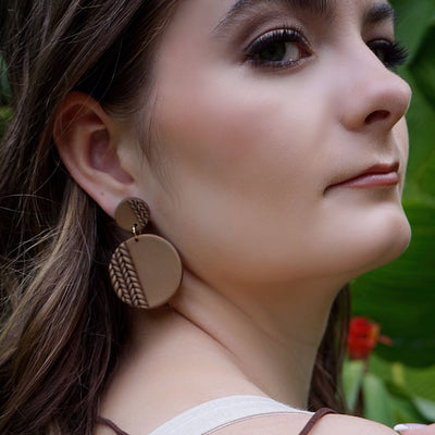 Woman wearing round clay earrings, surrounded by green foliage.