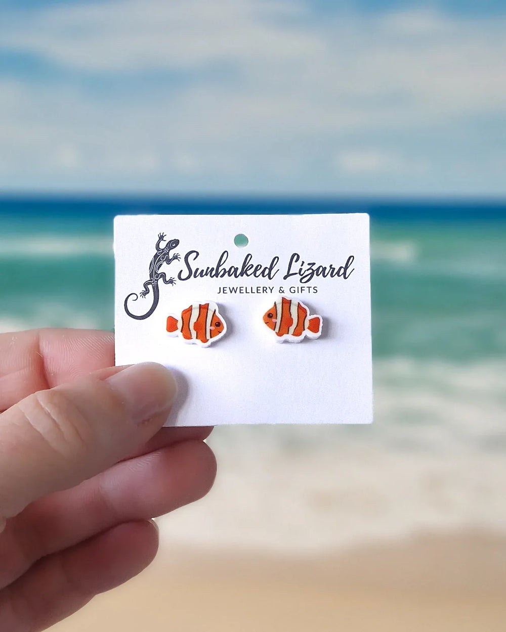 Card with clownfish earrings held by a hand against a beach background
