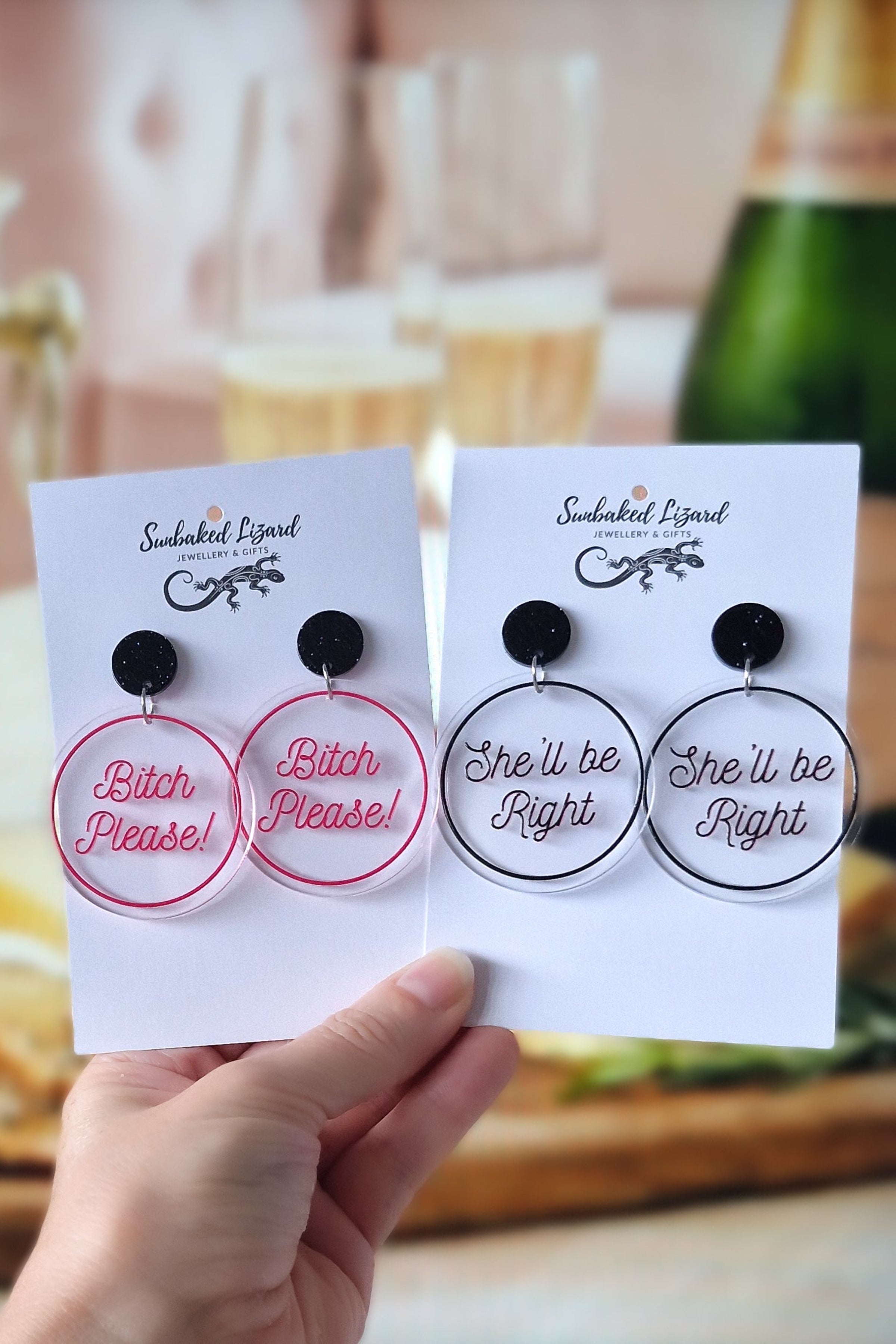 Two pairs of Sunbaked Lizard funny quote earrings on display cards, featuring Bitch Please and She'll Be Right designs with champagne in the background.