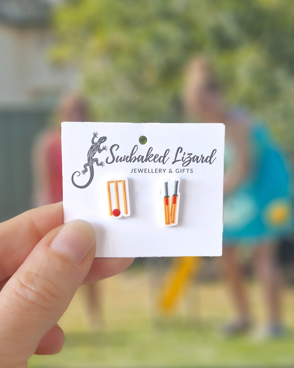 Cricket stud earrings featuring acrylic bat, ball and stumps on stainless steel posts on a white card, with a cricket game in the background