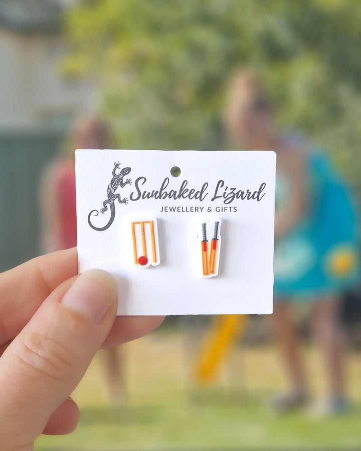 Cricket stud earrings featuring acrylic bat, ball and stumps on stainless steel posts on a white card, with a cricket game in the background