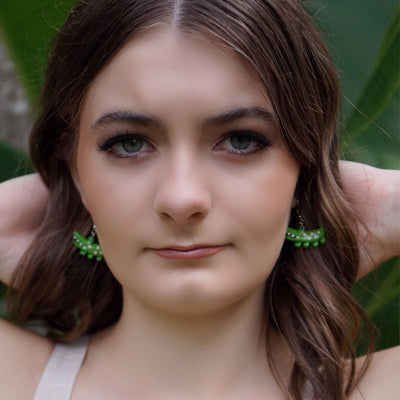 Woman with long brown hair wearing a light-colored apron against a green leafy background, wearing green crocodile earrings