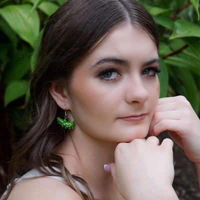 Woman with green crocodile earrings resting her chin n her hands against a leafy background