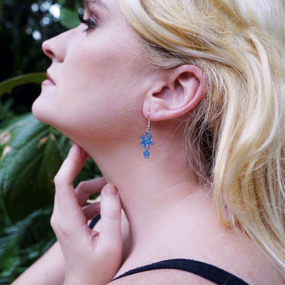 Close-up of a woman wearing blue flower pot earrings with a blurred leafy background