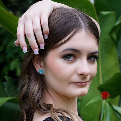 Woman wearing vibrant blue stud earrings with gold and opal flecks against a green leafy background