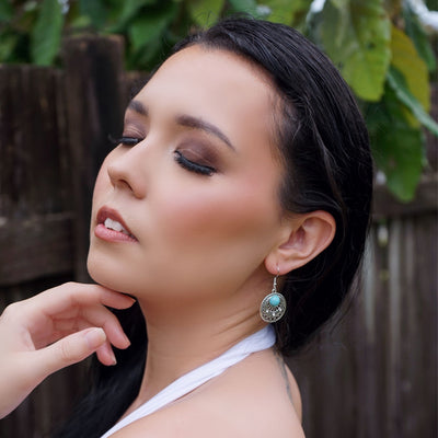 Woman with dark hair and earrings against a blurred fence and leaves background