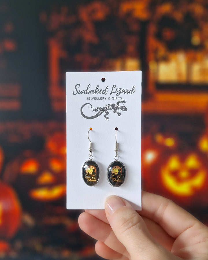 Hand holding glass oval Halloween earrings with black cat, yellow moon, bats and Happy Halloween text, on a jewellery backing card in front of a Halloween pumpkin background
