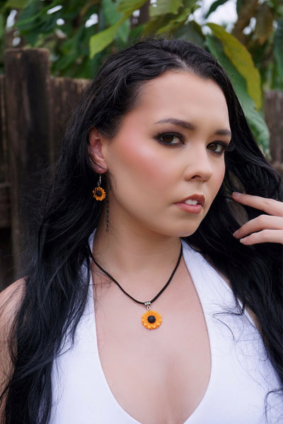 Woman with long dark hair and a necklace with a sunflower pendant outdoors.