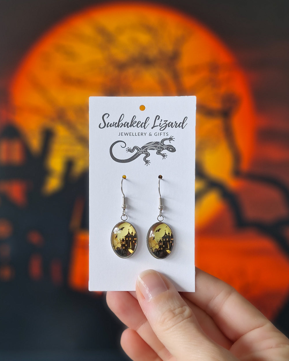 Hand holding glass oval haunted house earrings showing manor silhouette, bats and graveyard against amber moon, silver hooks with a Halloween background