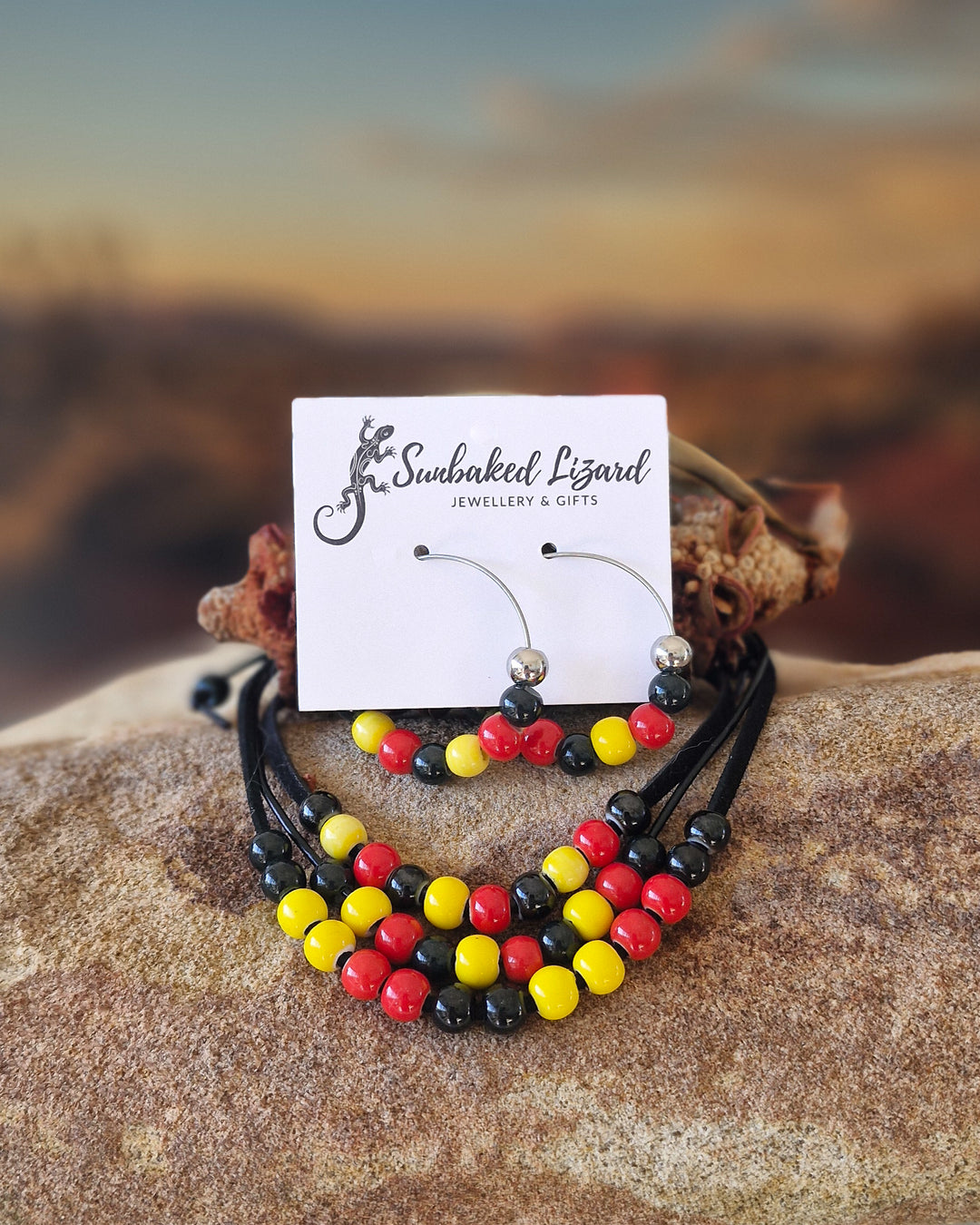 Collection of red, yellow and black Indigenous inspired jewellery with glossy beads on a sandstone rock with a blurred desert landscape in the background.