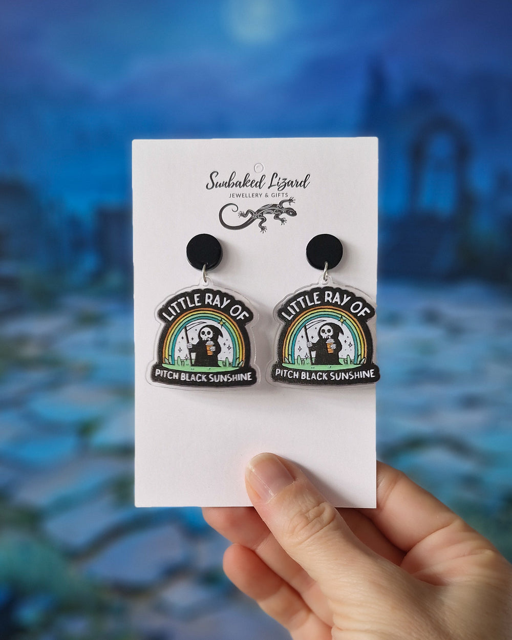 Hand holding Little Ray of Pitch Black Sunshine earrings with cartoon grim reaper holding iced coffee under rainbow