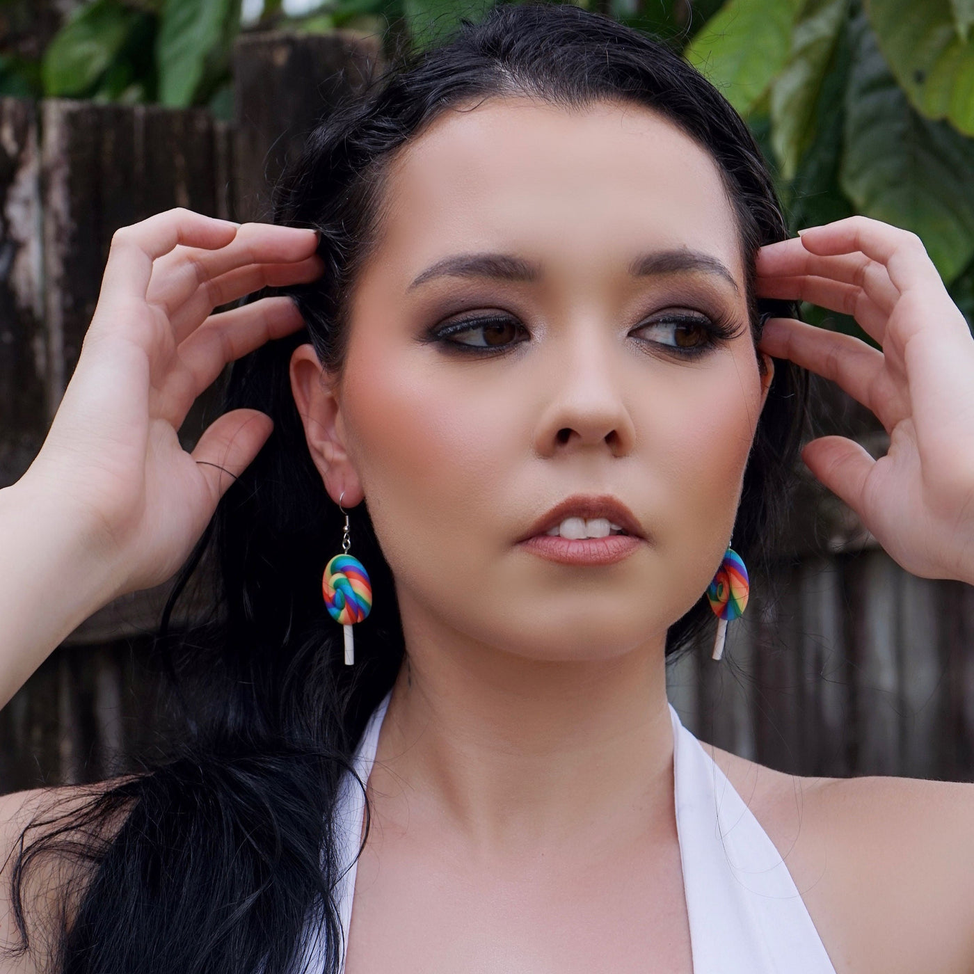 Woman with long dark hair and colorful lollipop earrings against a wooden fence