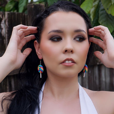 Woman with long dark hair and colorful lollipop earrings against a wooden fence