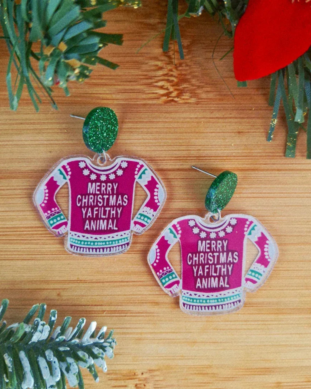 Pink sweater earrings with text on a wooden surface surrounded by Christmas decorations.