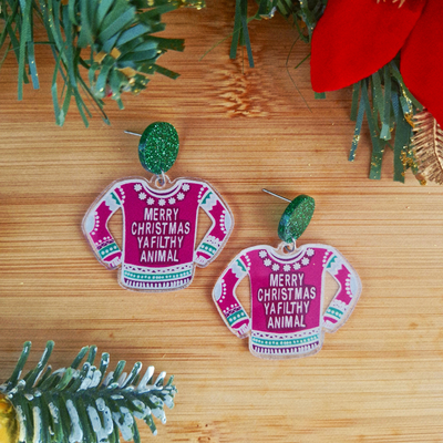 Pink sweater earrings with text on a wooden surface surrounded by Christmas decorations.