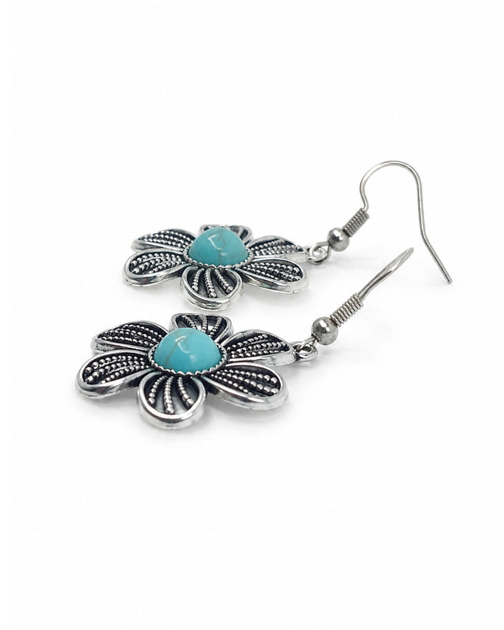 A pair of silver-toned dangle earrings shaped like flowers, featuring textured silver petals and a central turquoise-colored stone, shown on a plain white background.