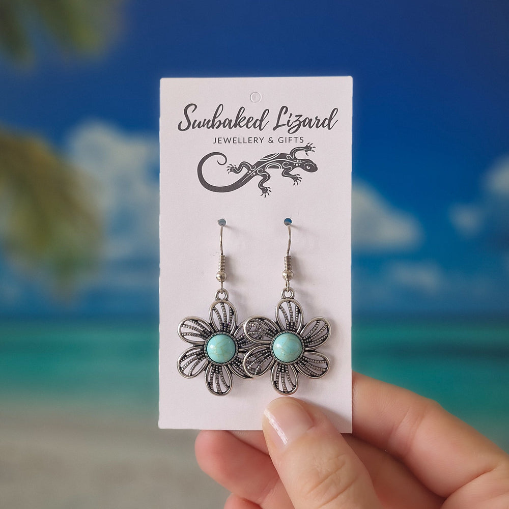A pair of silver filigree flower drop earrings with light blue-green turquoise centers, displayed on a white 'Sunbaked Lizard Jewellery & Gifts' card, held by a hand against a blurred beach background.